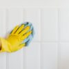 Crop anonymous housekeeper in yellow gloves cleaning white tiled wall using rag in bathroom