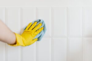 Crop anonymous housekeeper in yellow gloves cleaning white tiled wall using rag in bathroom