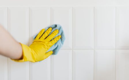 Crop anonymous housekeeper in yellow gloves cleaning white tiled wall using rag in bathroom